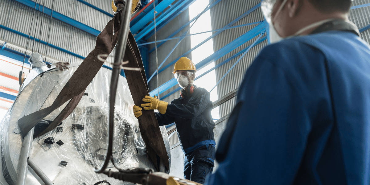 An industrial worker in a hard hat and face mask guides a large metal tank being lifted by an overhead crane, as a colleague observes.