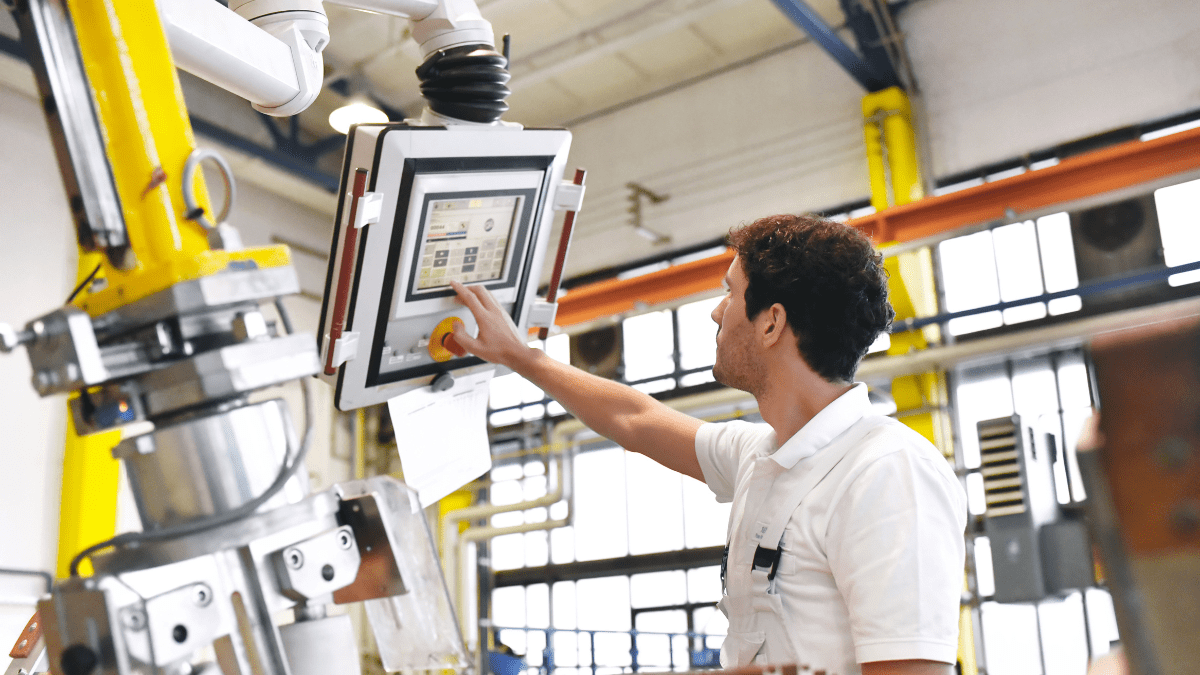 A male factory worker in a white shirt operates a modern robotic arm by using its attached digital touchscreen control panel in a factory setting.