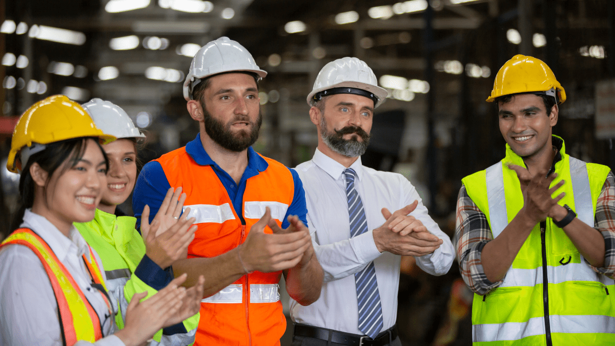 A diverse team of industrial workers and managers in hard hats and safety vests clapping and smiling to show appreciation on a factory floor.