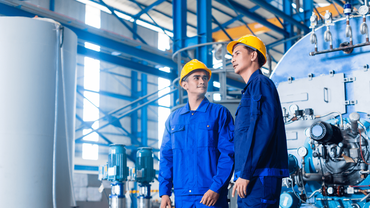 Two industrial workers in blue coveralls and yellow hard hats stand next to large blue machinery in a factory, looking up and discussing operations.
