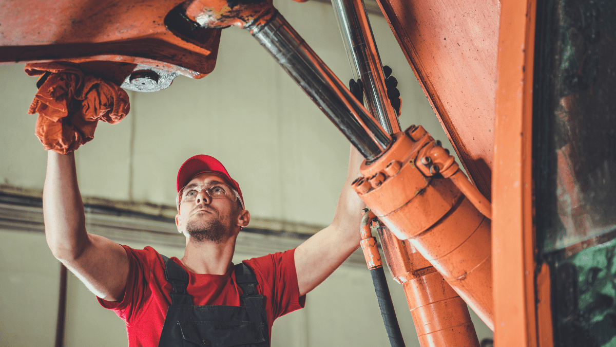 A male mechanic in a red cap and safety glasses looks up while performing maintenance on a large orange hydraulic machine in a workshop.