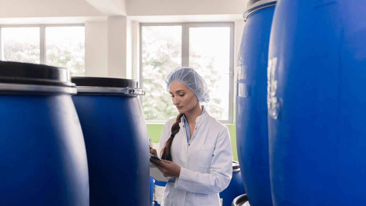A quality control inspector in a white lab coat and hairnet stands between large blue industrial barrels, writing notes on a clipboard.