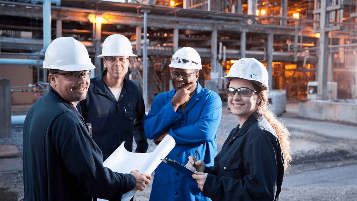 A diverse team of four industrial workers in white hard hats and safety glasses holding a safety meeting with blueprints and a clipboard at an industrial plant.