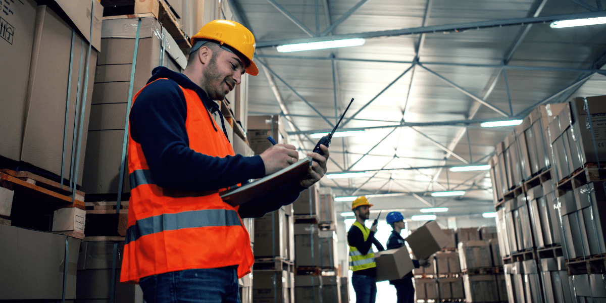 A warehouse supervisor in a hard hat and orange vest writes on a clipboard, holding a walkie-talkie, while other workers move boxes in the background.