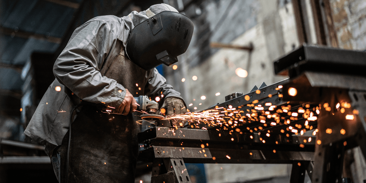A metalworker in a welding mask and gloves uses an angle grinder on a piece of metal, sending a bright shower of sparks in a workshop.