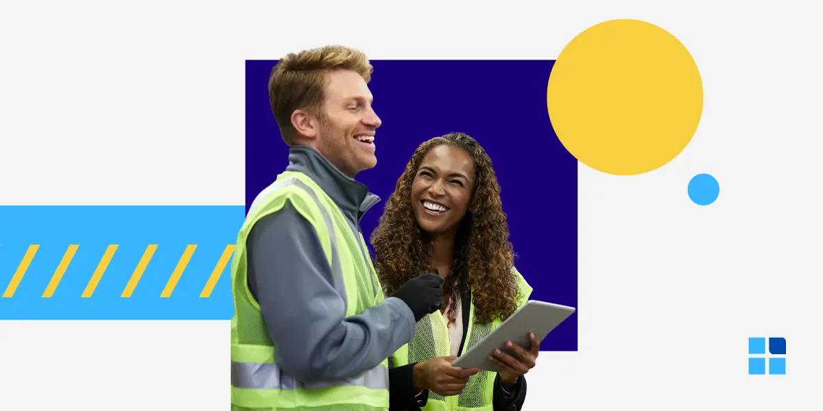 Two people in high-visibility vests are smiling and interacting while holding a tablet.