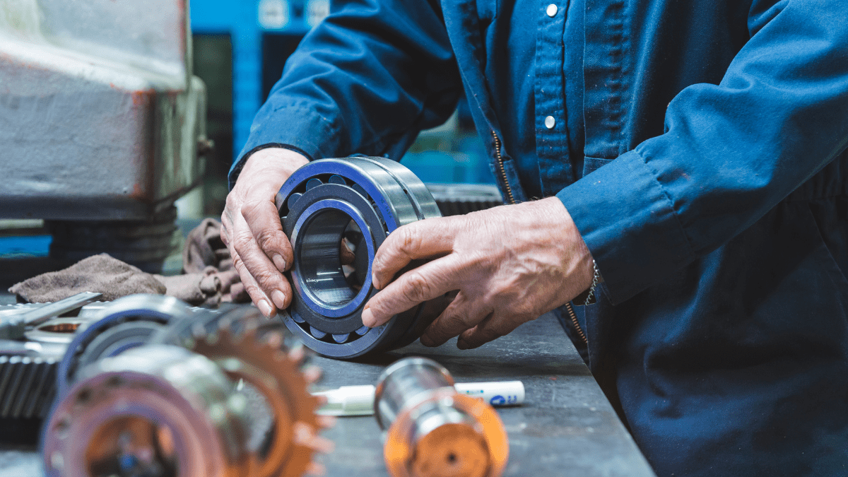 Close-up of a factory worker's hands in blue coveralls, performing industrial maintenance on a large machine bearing on a workbench.