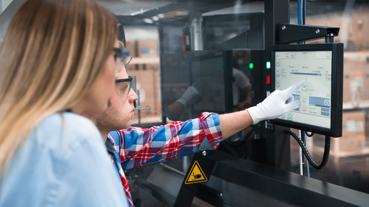 A male factory worker in a plaid shirt and glove points to a digital touchscreen control panel on an industrial machine, as a female colleague observes.