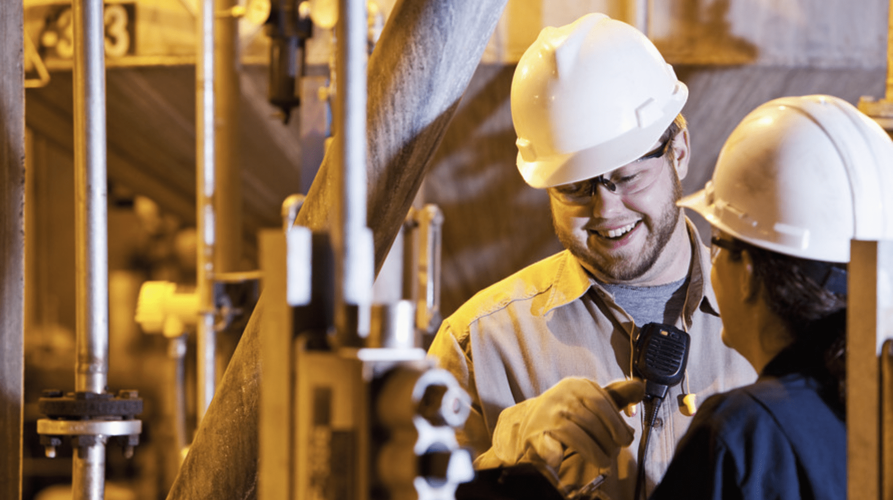 Two industrial workers in white hard hats surrounded by pipes, where the smiling man talks into a walkie-talkie to his female colleague.