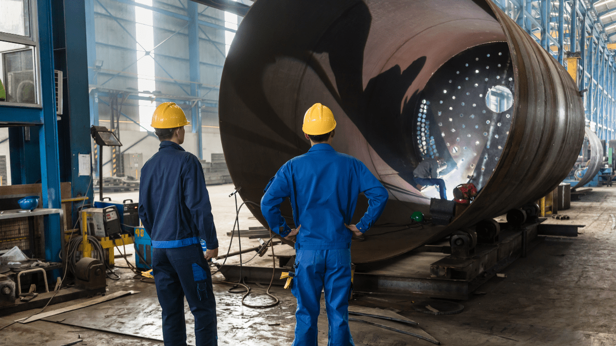 Three industrial workers in a large fabrication plant, where two observe a third worker welding inside a massive cylindrical metal vessel.