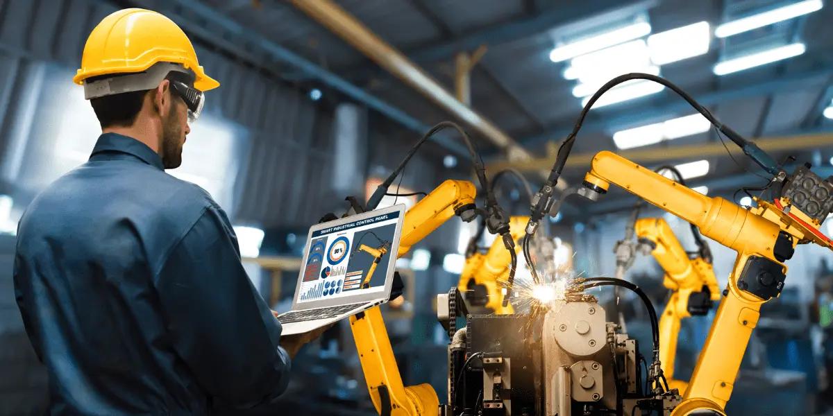 A worker in a yellow hard hat and safety glasses uses a laptop to monitor robotic arms.