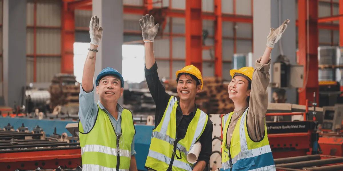 Three workers in a factory, wearing helmets and reflective vests, smiling and waving.