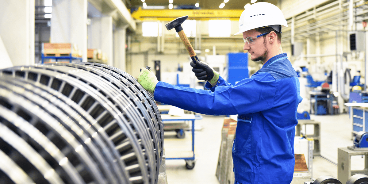 An industrial worker in a hard hat and safety glasses uses a hammer to perform maintenance on a large metal turbine in a factory.