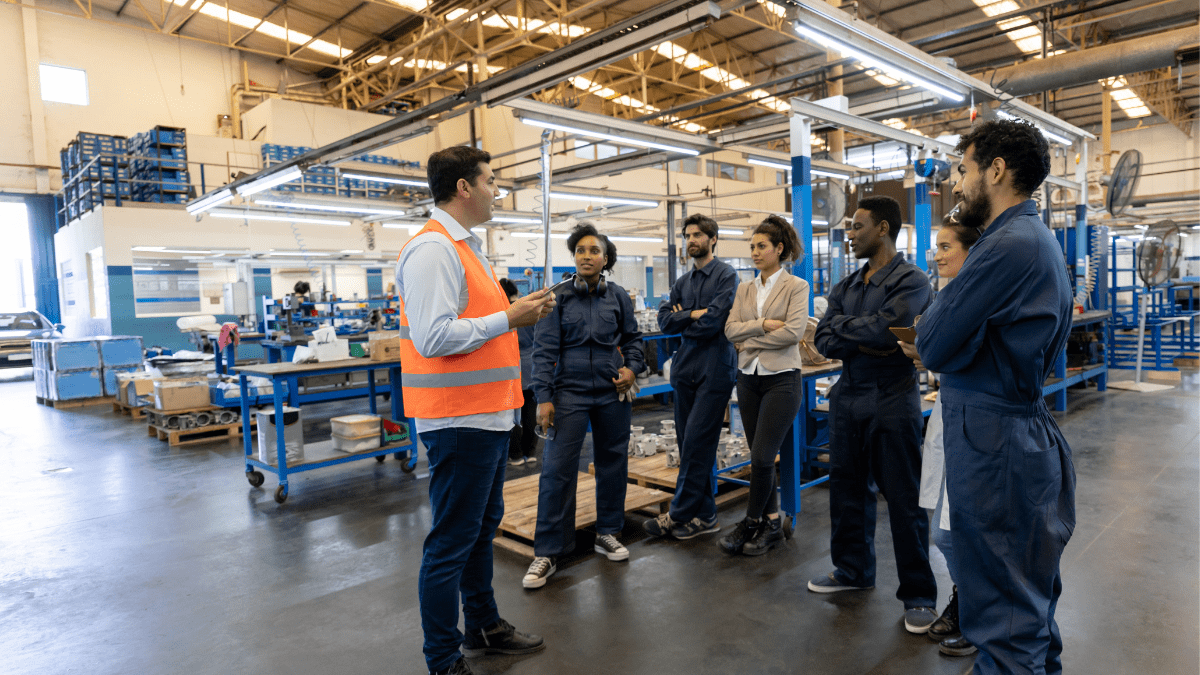 A manager in an orange safety vest leads a team briefing for a diverse group of six workers on a spacious factory floor.