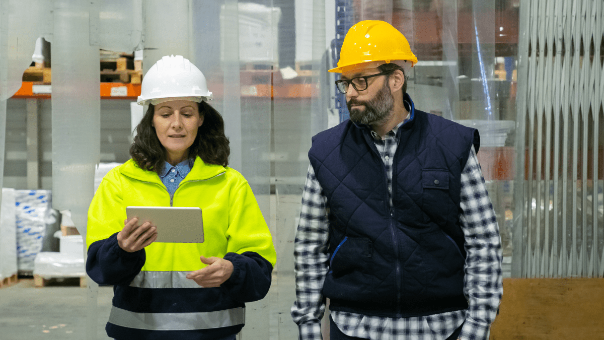 A female worker in a white hard hat and high-vis jacket holds a tablet while walking and talking with a male worker in a yellow hard hat and vest in a warehouse.