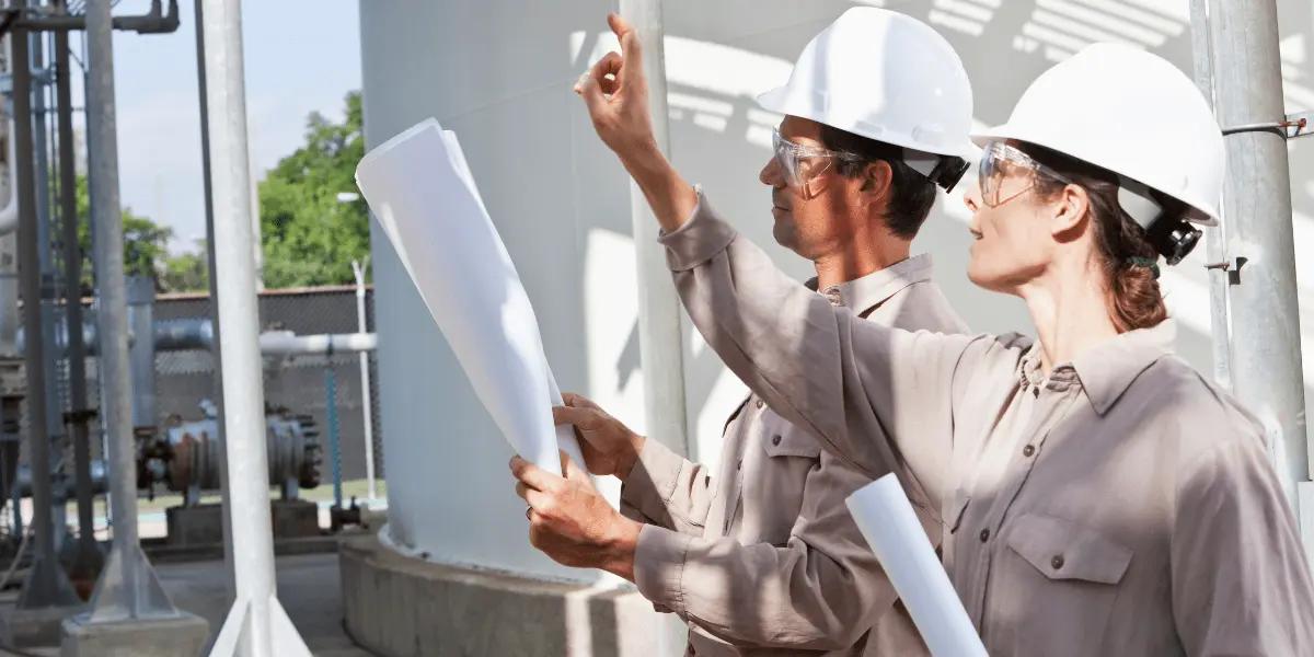 Two engineers wearing white hard hats and safety glasses examine blueprints at an industrial site.