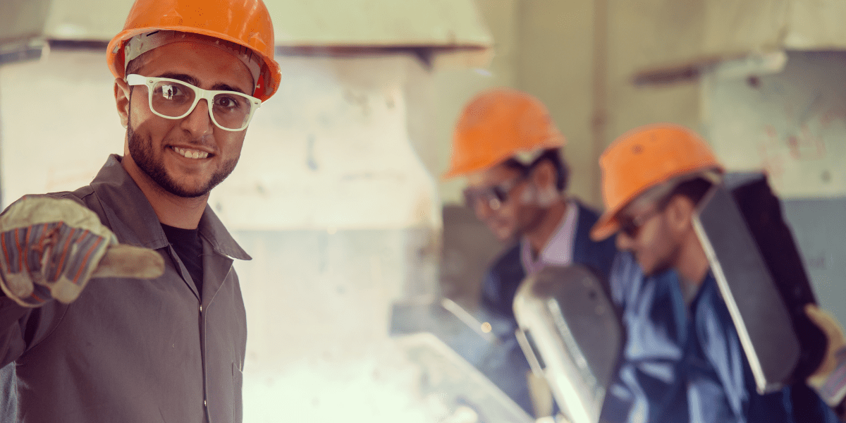 A smiling industrial worker in a hard hat and safety glasses points at the camera, while two other workers weld in the factory background.