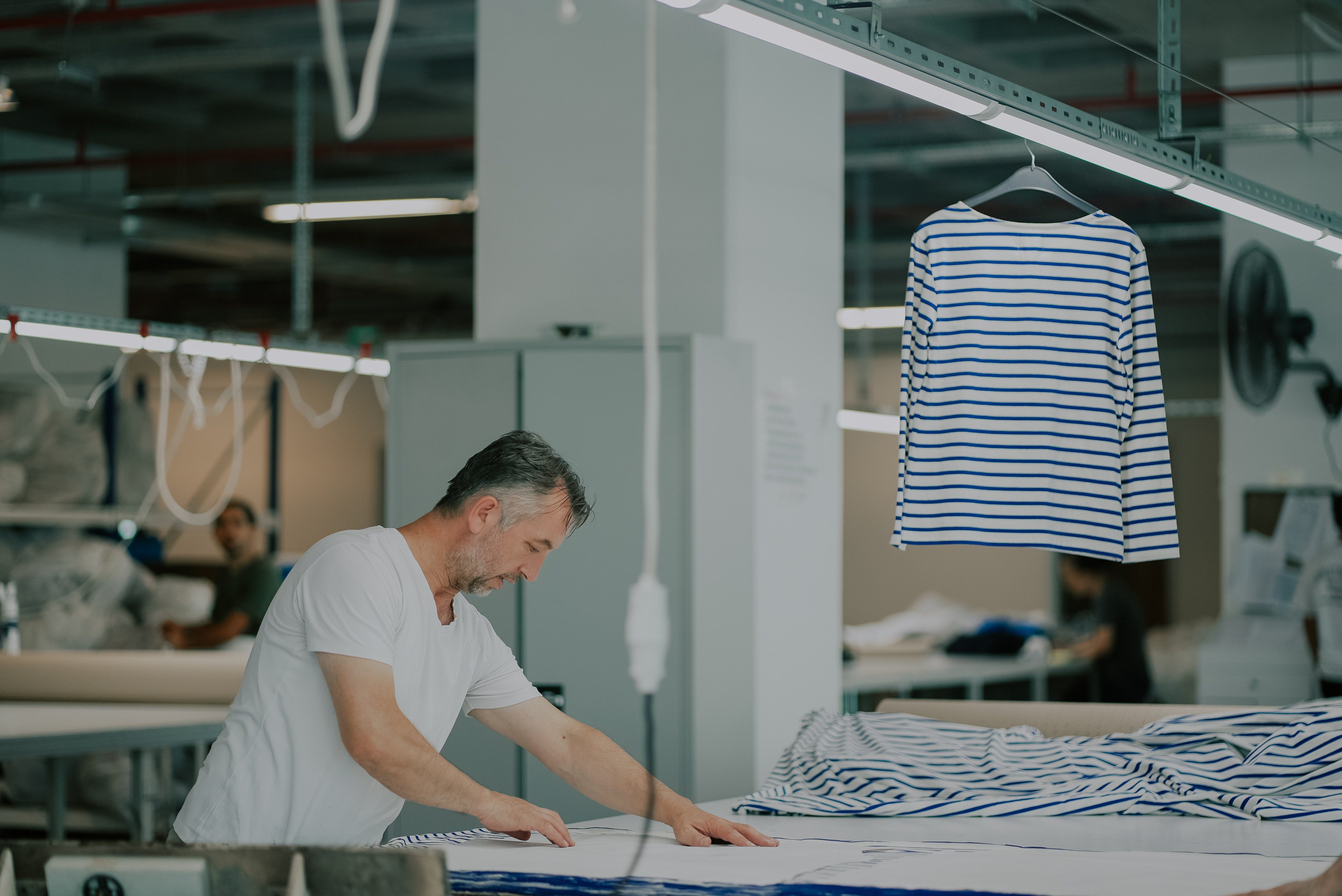 A male worker in a white t-shirt leans over a large table in a garment factory, laying out a pattern on blue and white striped fabric.