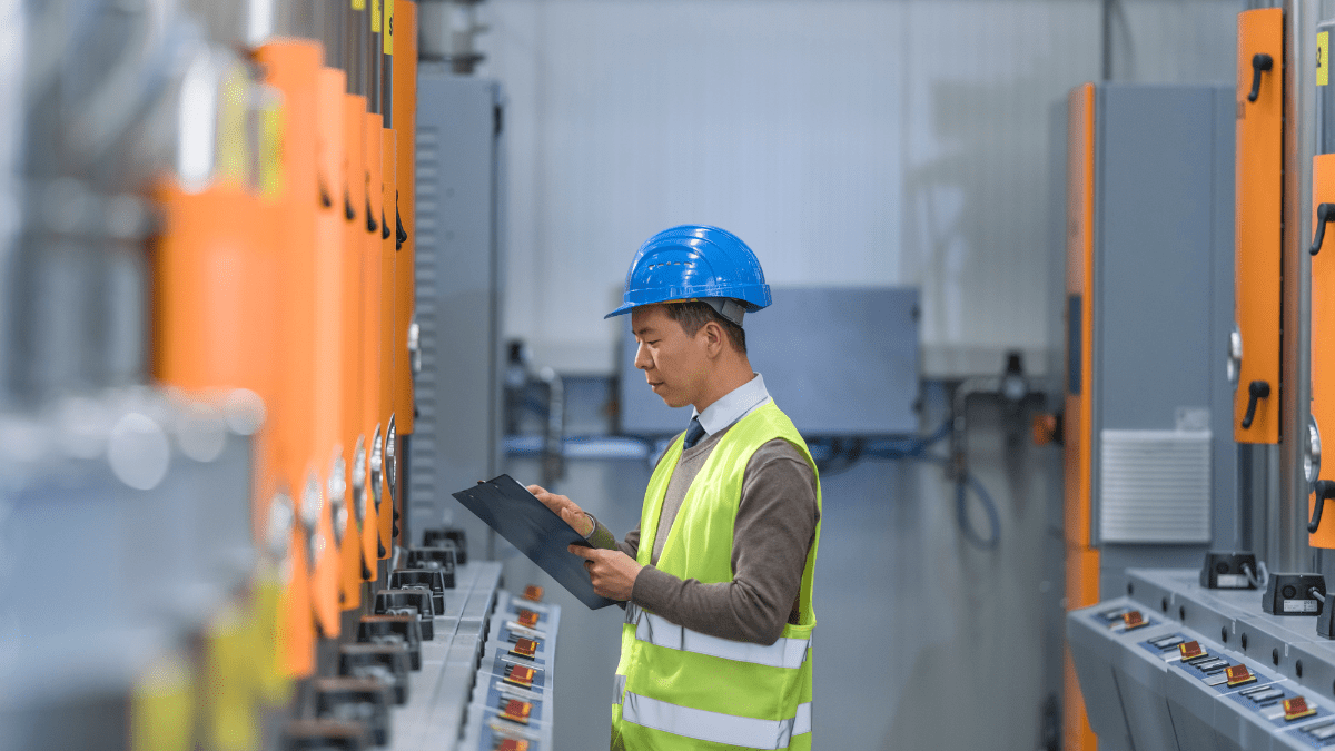 An industrial engineer in a blue hard hat and yellow vest holds a clipboard while inspecting a row of orange and grey machinery in a factory.