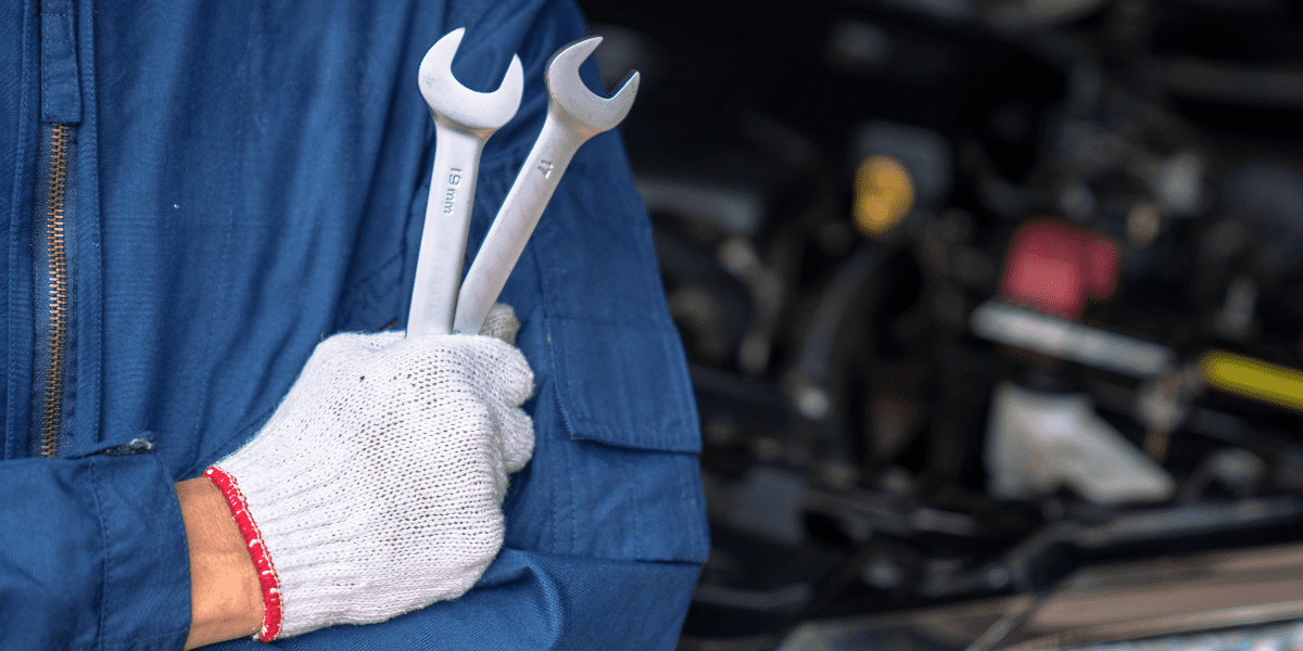 Close-up of a mechanic in blue coveralls and a white glove, holding two wrenches with arms crossed in front of a car engine.