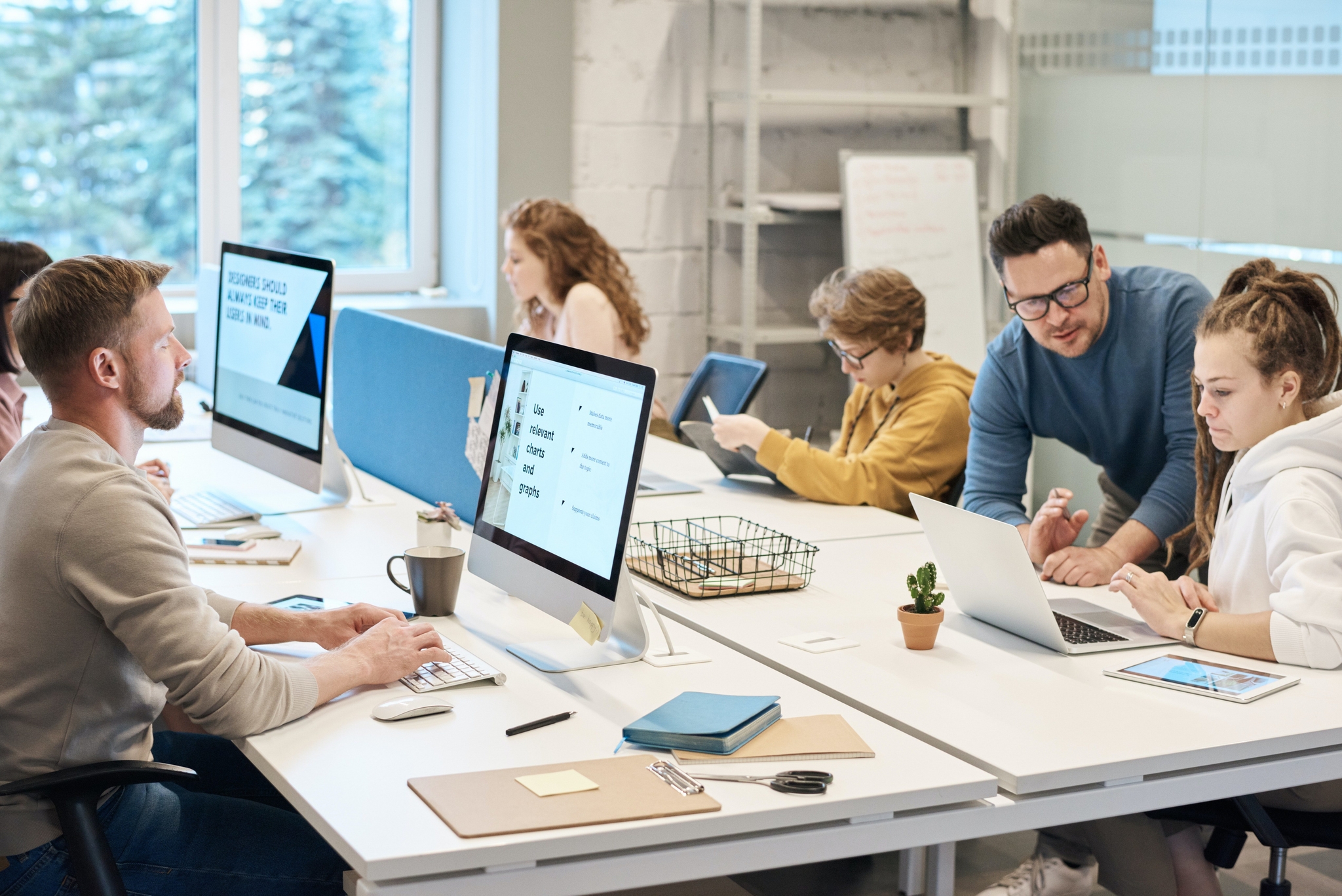 A busy, modern open-plan office with a team of colleagues working at desks, including a man helping a woman at a laptop.