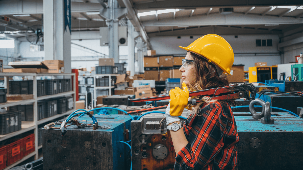 A female industrial worker in a yellow hard hat and safety goggles holds a large pipe wrench on her shoulder while standing on a factory floor.