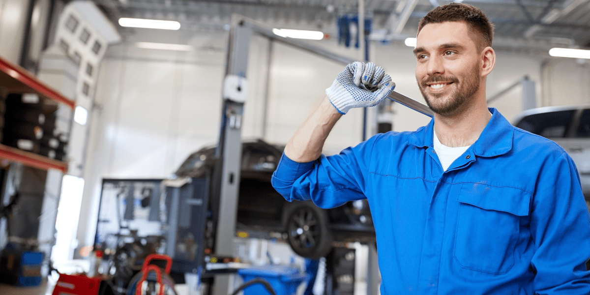 A smiling auto mechanic in blue coveralls holds a wrench on his shoulder, standing in a garage with a car on a lift.