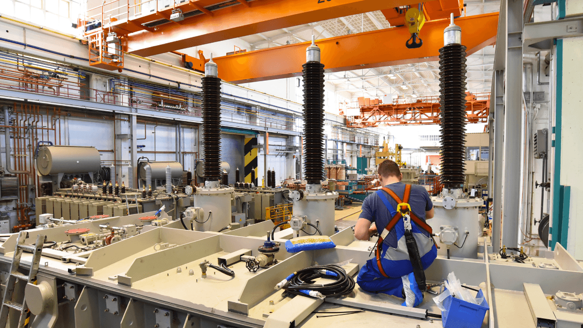 A technician in a safety harness works on a large electrical transformer in a heavy manufacturing plant with orange overhead cranes.