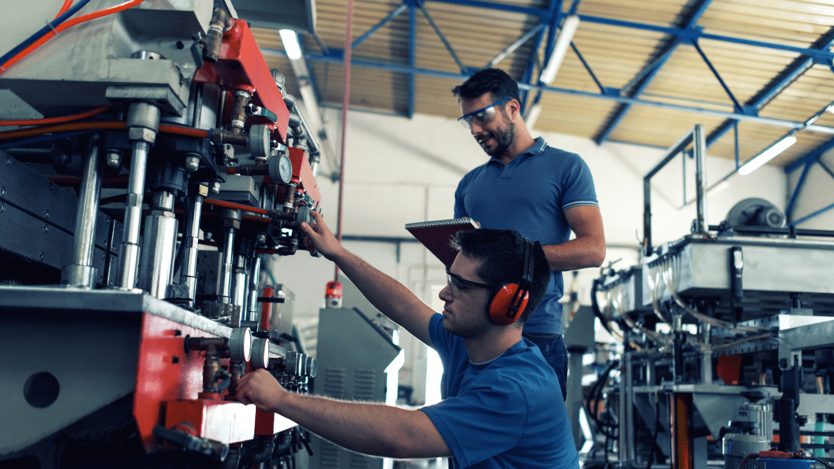 A factory worker in earmuffs and safety glasses adjusts a large industrial machine while a supervisor with a clipboard observes and takes notes.